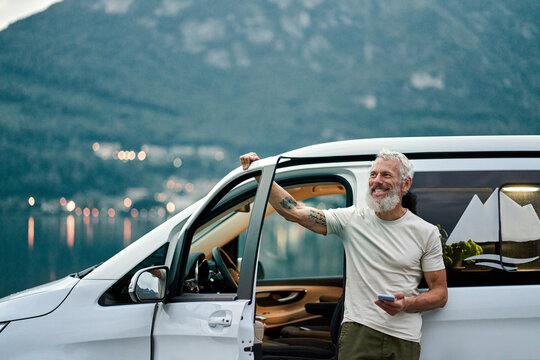 Happy older man standing near rv camper van on vacation using mobile phone. Smiling mature active traveler holding smartphone enjoying free roaming in camping tourism in nature park feeling freedom. - Powered by Adobe