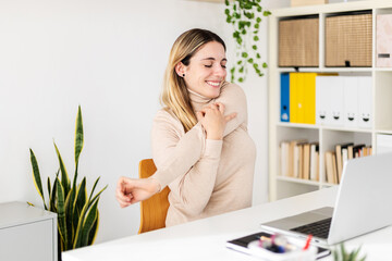 Young adult woman sitting on home office table stretching arms while working on laptop computer. Freelancer female worker taking break from work. Health concept - obrazy, fototapety, plakaty