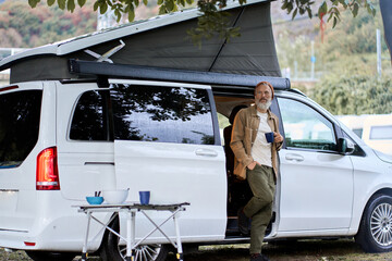 Active older hipster man standing near rv camper van on vacation trip. Mature traveler looking away...