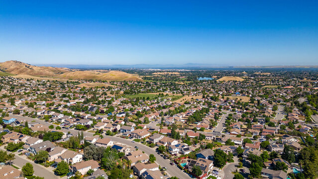 .A breathtaking aerial view of Antioch, California: houses nestled amidst verdant hills, crisscrossed by meandering streets, all under a picturesque blue sky.