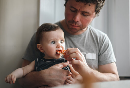 A caucasian baby girl sits in the arms of her dad, who gives her a piece of homemade pizza