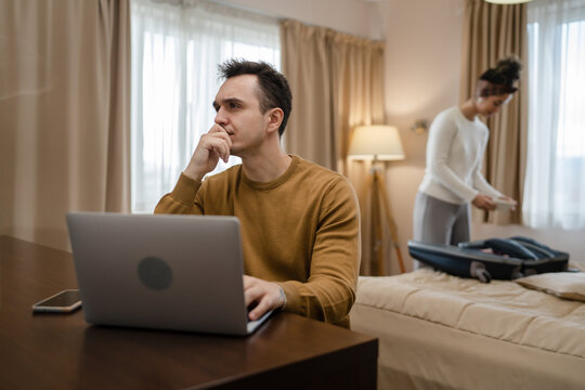 couple family life caucasian man working on laptop computer or making online reservations while his wife young woman is packing or unpack baggage suitcase on bed in hotel room real people copy space