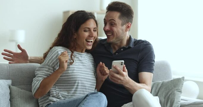 Happy couple sit on sofa staring at cellphone screen, scream with joy, hugging white get fantastic commercial offer, reading great online news, amazed by win in interactive game, get prize in lottery