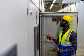 Naklejka na ściany i meble Professional African male engineer in safety uniform working at factory server electric control panel room. Industrial technician worker maintenance checking power system at manufacturing plant room.