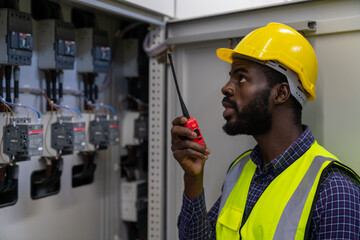 Professional African male engineer in safety uniform working at factory server electric control panel room. Industrial technician worker maintenance checking power system at manufacturing plant room.