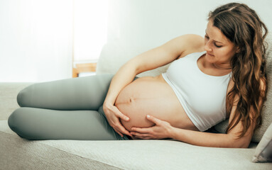 View of woman lying on her side on a sofa proudly holding her belly in the last stage of pregnancy. Pregnancy third trimester - week 34. Side view. White flat background.