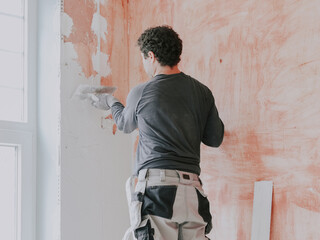 A young caucasian guy applies fresh putty with a small spatula on the wall near the window