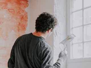 Caucasian young plasterer guy looks at the window opening for applying fresh putty with a spatula