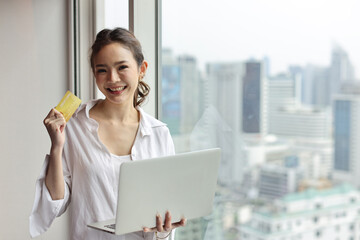 Asian woman in casual standing and holding credit card and using computer in house living room for online shopping payment. Confident businesswoman with city background. Lifestyle technology concept