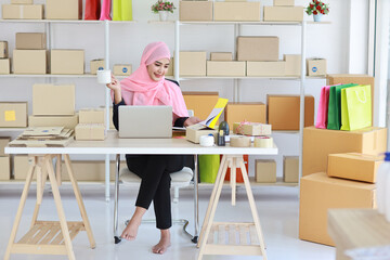 Active smiling asian woman in blue muslim suit sitting and working with online package box delivery. Startup small business SME freelance girl working on computer and mobile phone with happy face.