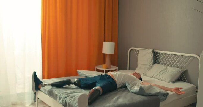 happy cheerful disabled man with artifical legs falls down with his arms outstretched on a soft cozy bed, lies and rests in slow motion Never give up new life strength man having rest