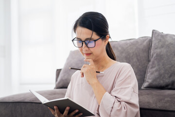 Mature asian woman wearing glasses taking notes in living room at home.