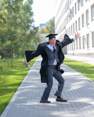 Old happy man in graduation gown jumping outdoors and holding diploma. Vertical.