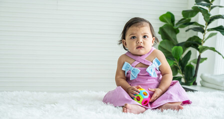 Fototapeta premium Adorable mixed race baby girl in pink clothes sitting on bed at home, looking away, playing toy color ball. Kid at home and childhood concept