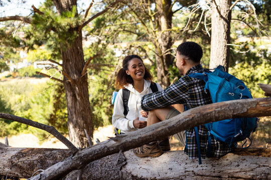 Happy biracial couple wearing backpacks sitting on tree trunk in forest