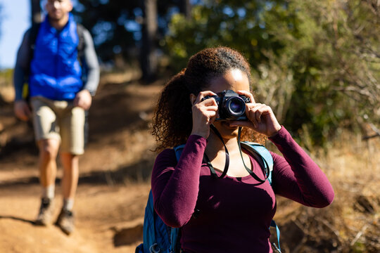 Happy biracial couple wearing backpacks, hiking and taking pictures with camera in forest