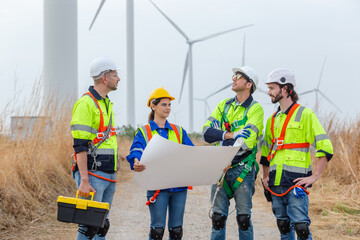 Teamwork engineer worker wearing safety uniform holding and reading blueprint at wind turbine field...