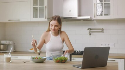 The girl, dressed in blue jeans and a white shirt, is preparing a salad in the kitchen. The woman tosses a salad for herself and watch tv series on laptop