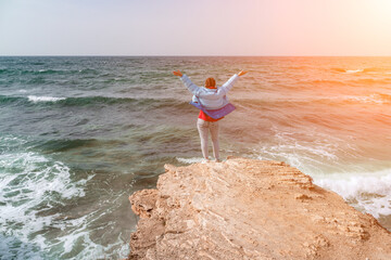 A woman in a blue jacket stands on a rock above a cliff above th