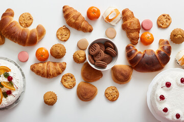 Top view of table with various cookies, cakes, croissants, bagels on white background.