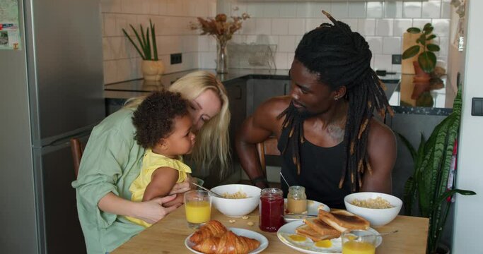 little adorable cute African daughter sitting on mom's knees Slow motion Mother gives juce, food to daughter. beautiful black child is drinking fresh juice on breakfast family at home in cozy kitchen.