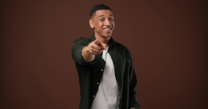 Playful african american guy pointing at camera and smiling standing over brown studio background