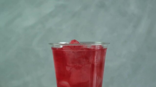 Ice cubes falls into disposable plastic cup of red fruit ice tea, close-up. Slow motion. Refreshing summer drink concept. Street food, takeaway drinks.