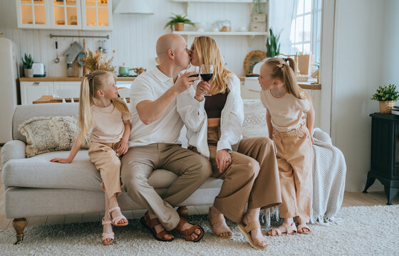 Caucasian family celebrating new home sitting on couch. Bald young man kissing wife holds glass of wine. Parents with daughters at home.