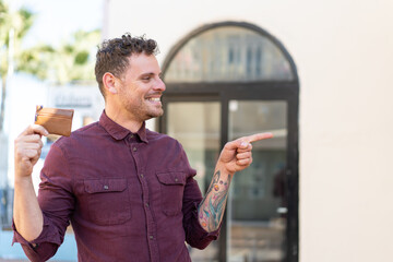 Young caucasian man holding a wallet at outdoors pointing to the side to present a product