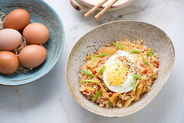 Bowl of kimchi fried rice topped with fried chicken egg on a light-beige marble background, high angle view, horizontal shot