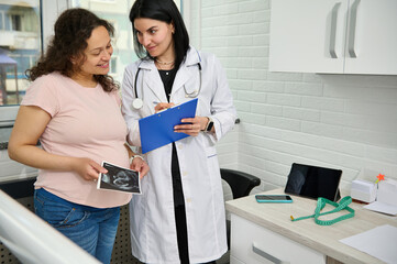 Obraz premium Multi ethnic happy positive pregnant woman holding the ultrasound scan of her baby, smiling while reading the prescription of her doctor gynecologist obstetrician during gynecological appointment