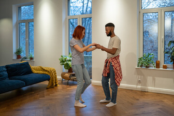 Young diverse couple in love preparing first wedding dance together, black man and caucasian girl dancing in living room, African guy teaching girlfriend how to dance during home date night