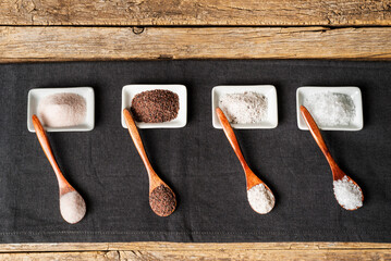 Wooden spoons next to some small plates with different types of salt, black, pink, flakes and seasoned with truffle, on a tablecloth on a rustic wooden background.Top view.