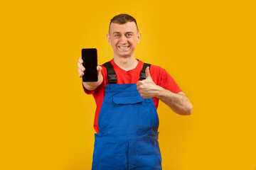 Young man in blue jumpsuit and red T-shirt holds smartphone with blank black screen and shows thumbs up. Copy space, mock up.