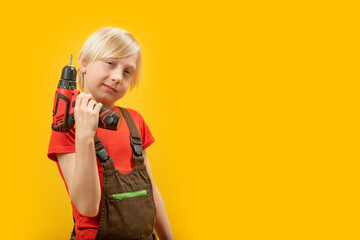 Studio portrait of blond boy in work clothes with drill in his hands in isolation on yellow background. Copy space.