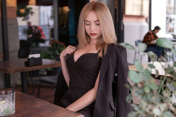 Sexy young blonde woman in black suit sits in cafe waiting for an order. Modern businesswoman.
