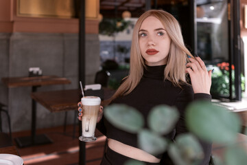 Pensive attractive blonde woman sits in coffee shop with cup of cappuccino coffee latte. beautiful girl alone in cafe.
