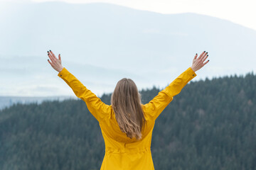 Girl wears yellow jacket raised hands and looks at mountains. Back view on nature background. Tourism and freedom - obrazy, fototapety, plakaty