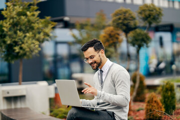 A young happy businessman in smart casual is sitting outside in park and typing on a laptop while smiling at it.