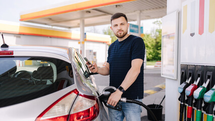 Bearded man refuelling car on gas station and looking into his smartphone. Man compares fuel prices 