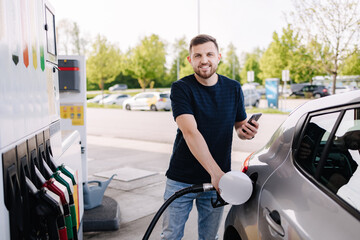 Bearded man refuelling car on gas station and looking into his smartphone. Man compares fuel prices 