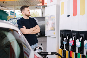 Handsome bearded man cross the arms during refueling car. Male looking on the scoreboard while...
