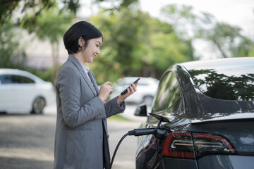 Asian female charging electric car parked in the nature area and adjusting an EV charging app on a...
