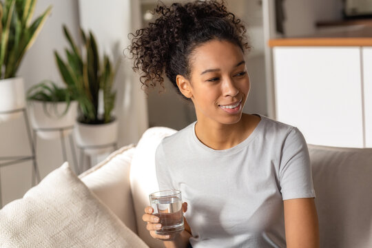 Beautiful African American woman holding glass of clean fresh water sitting on the sofa, relaxation at home, looking away and smiles. Healthy lifestyle