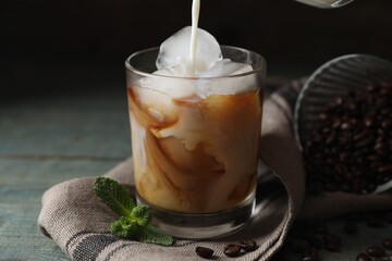Pouring milk into glass of delicious iced coffee, beans and mint on light blue table, closeup