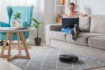 Robotic vacuum cleaner cleaning carpet, woman using laptop sitting on sofa at home