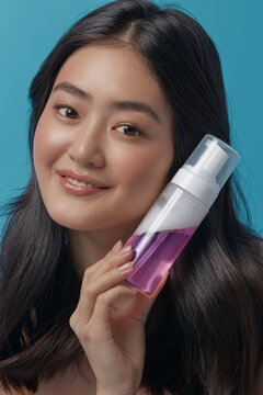 Beautiful korean ethnicity woman with a clean healthy skin holding near her face pink micellar water for facials cleansing and moisturizing after make up over blue background, close up vertical shot.