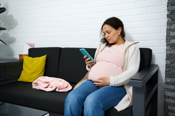 Multi ethnic adult pregnant woman checking her mobile phone while sitting on the sofa in the...