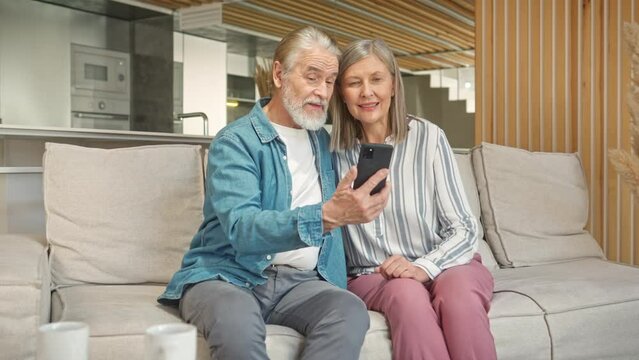 Handsome mature man showing family photo his woman on smartphone while sitting on cozy sofa. Happy from joyful memories of past. Actively discussing events which seeing on frame. Resting together.