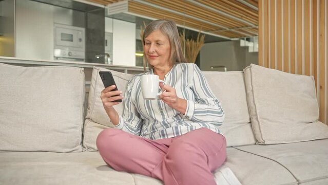 Zooming in on Caucasian woman with gray hair relaxing on soft couch in modern room. Beautiful mature female holding cup of hot drink while using cellphone. Leisure day at home. Concept of weekend.